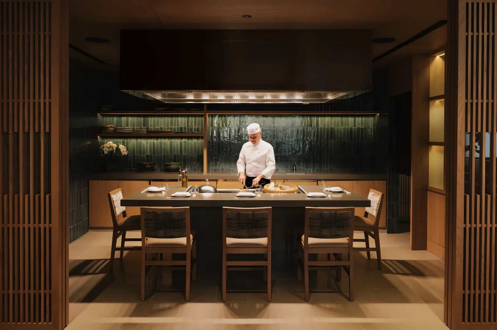 A chef in a white uniform stands at a modern kitchen island, preparing food. The room is warmly lit with wooden decor, giving a calm, inviting atmosphere.