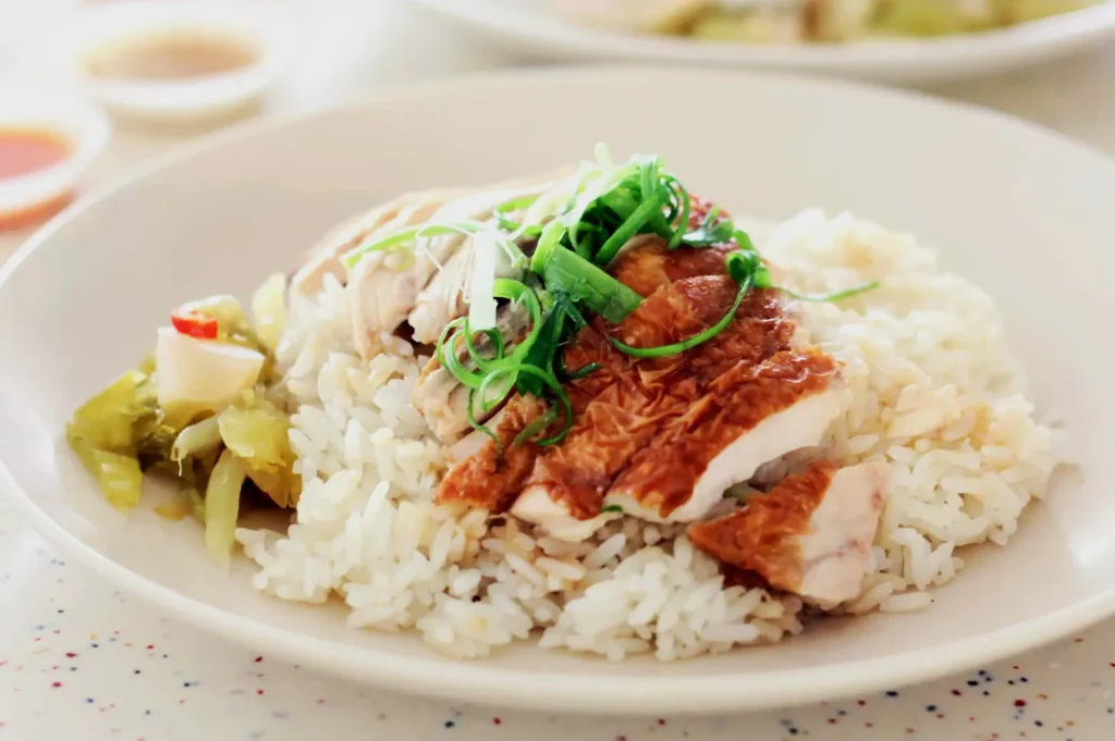 A plate of Hainanese chicken rice topped with sliced roasted chicken, garnished with vibrant green onions. A side of pickled vegetables adds color.