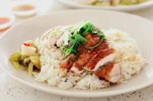 A plate of Hainanese chicken rice topped with sliced roasted chicken, garnished with vibrant green onions. A side of pickled vegetables adds color.