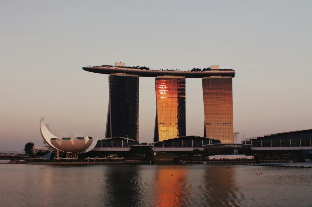 Singapore's Marina Bay Sands with a sunset reflection on its facade, flanked by the ArtScience Museum. The scene is calm, mirrored in still waters.