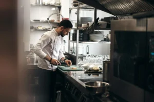 A focused chef in a white uniform chops vegetables in a professional kitchen, surrounded by stainless steel appliances and neatly stacked dishes.