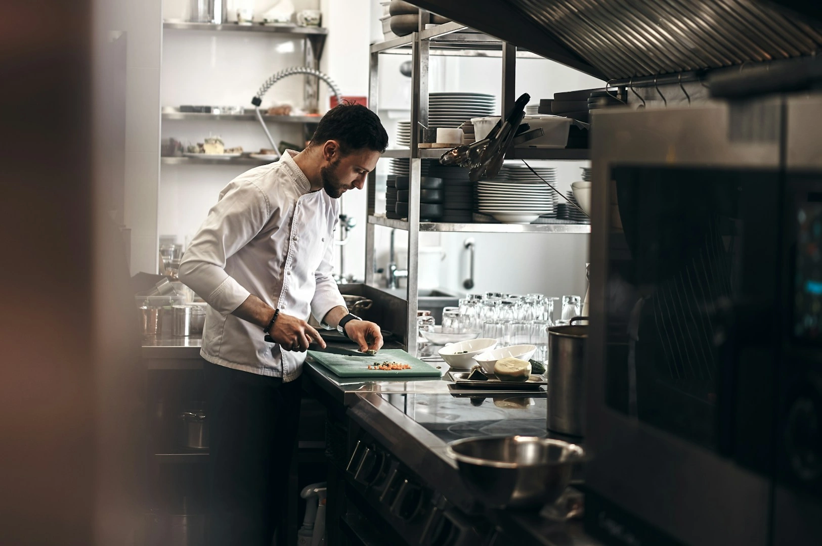A focused chef in a white uniform chops vegetables in a professional kitchen, surrounded by stainless steel appliances and neatly stacked dishes.