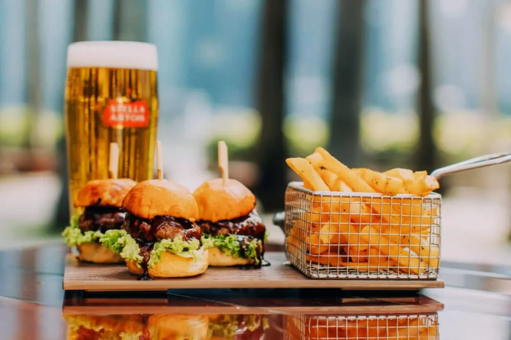 Sliders on a wooden board with fresh lettuce and sauce, beside a basket of golden fries, and a tall glass of beer in a sunlit outdoor setting