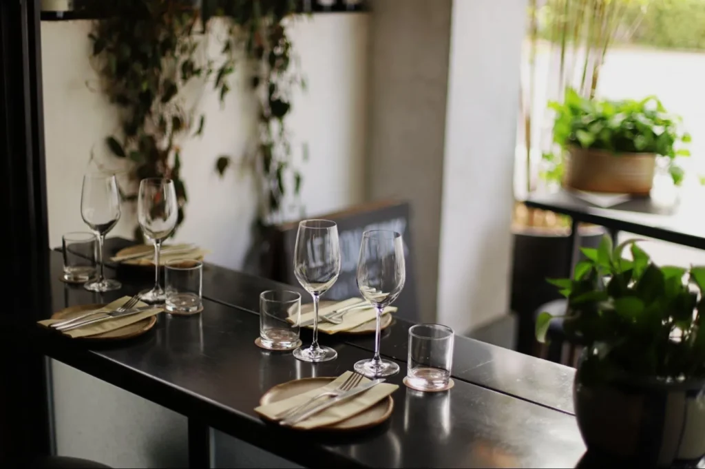 Elegant restaurant table set with glassware, plates, and folded napkins, surrounded by lush green plants, creating a cozy, intimate ambiance.