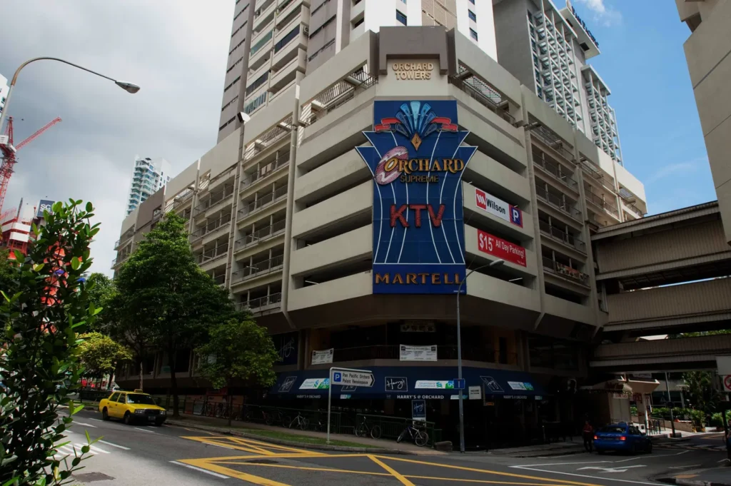 Street view of Orchard Towers, a multi-story building with a prominent "KTV" sign, surrounded by lush trees and city traffic under a partly cloudy sky.