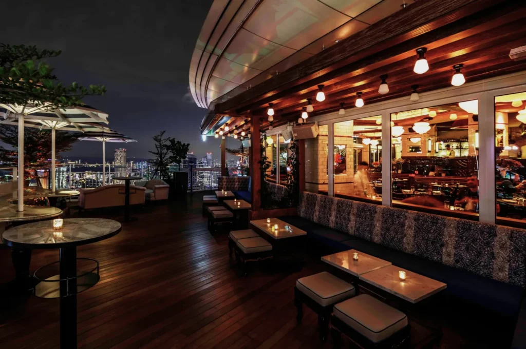 Stylish rooftop bar at night with wooden flooring, cozy seating, and city skyline views. Soft lighting creates an inviting and elegant atmosphere.