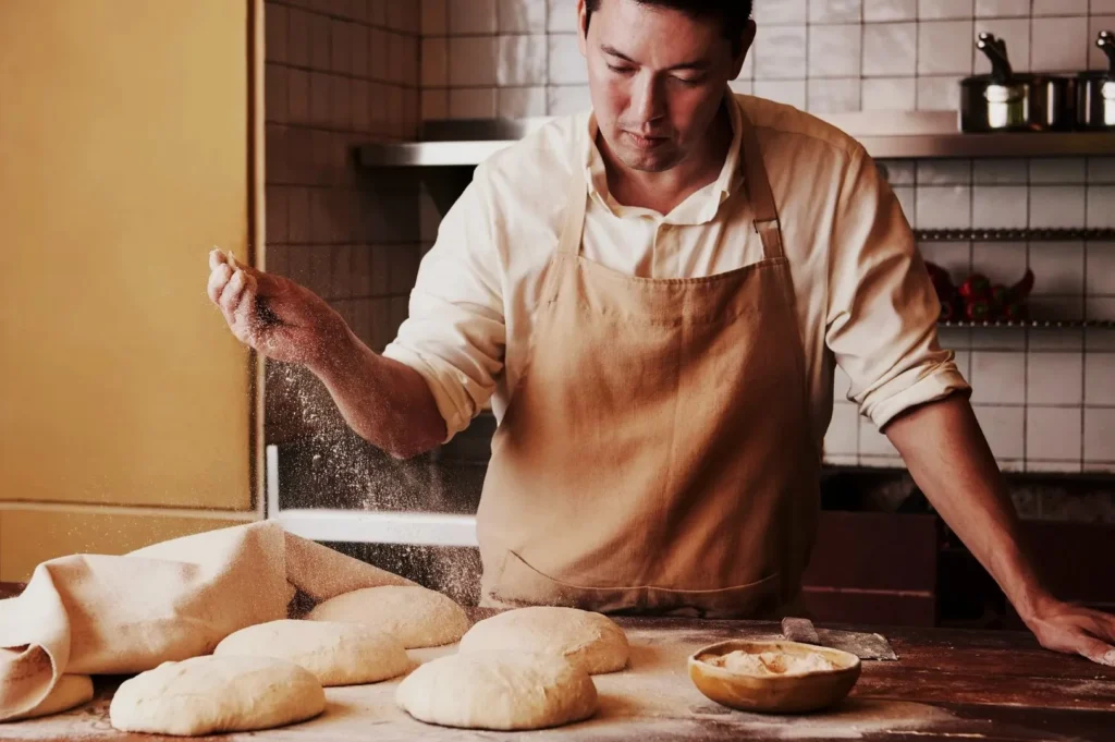 Baker in an apron sprinkles flour over dough in a warm kitchen, creating a focused, artisanal atmosphere with bread-making essentials nearby.