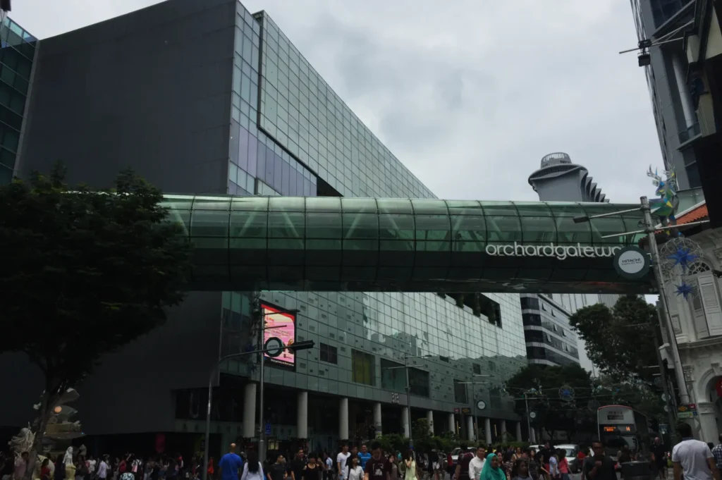 Orchard Gateway's glass bridge against a modern cityscape, bustling with people. Overcast sky enhances the vibrant urban scene.