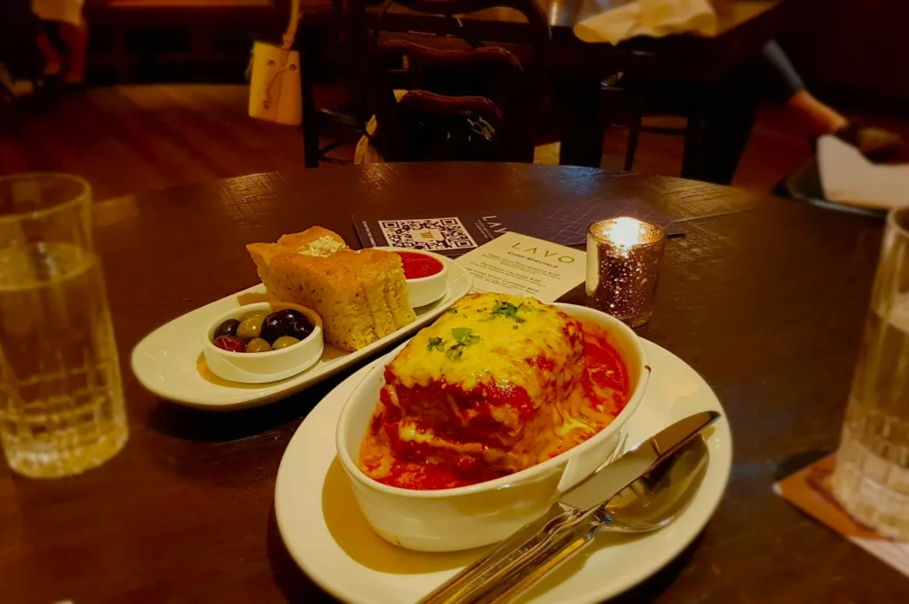 A cozy restaurant setting with a candlelit table showcasing a steaming dish of lasagna, accompanied by bread, olives, and a glass of water.