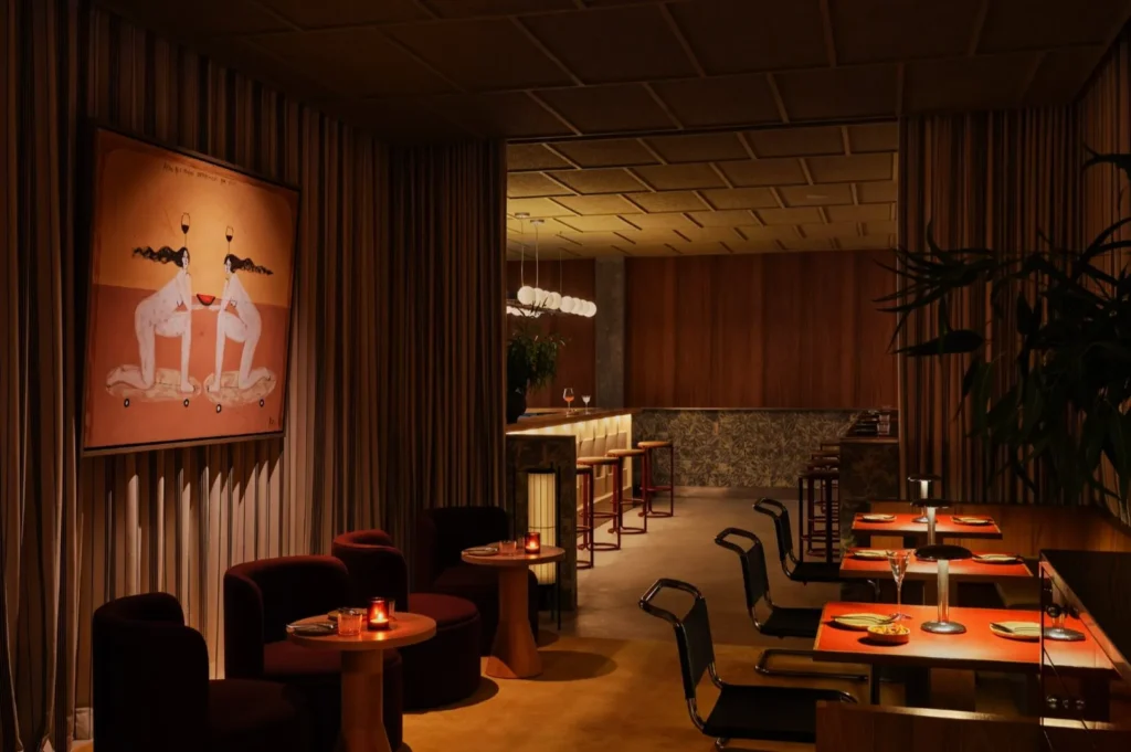 Warmly lit dining area with modern decor, featuring orange tables, black chairs, bar stools, and a large, whimsical painting on a curtain-draped wall. Cozy atmosphere.