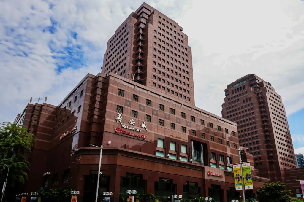 A large, modern, multi-story building with a red brick facade labeled as "Ngee Ann City," featuring banners, greenery, and a partly cloudy sky.