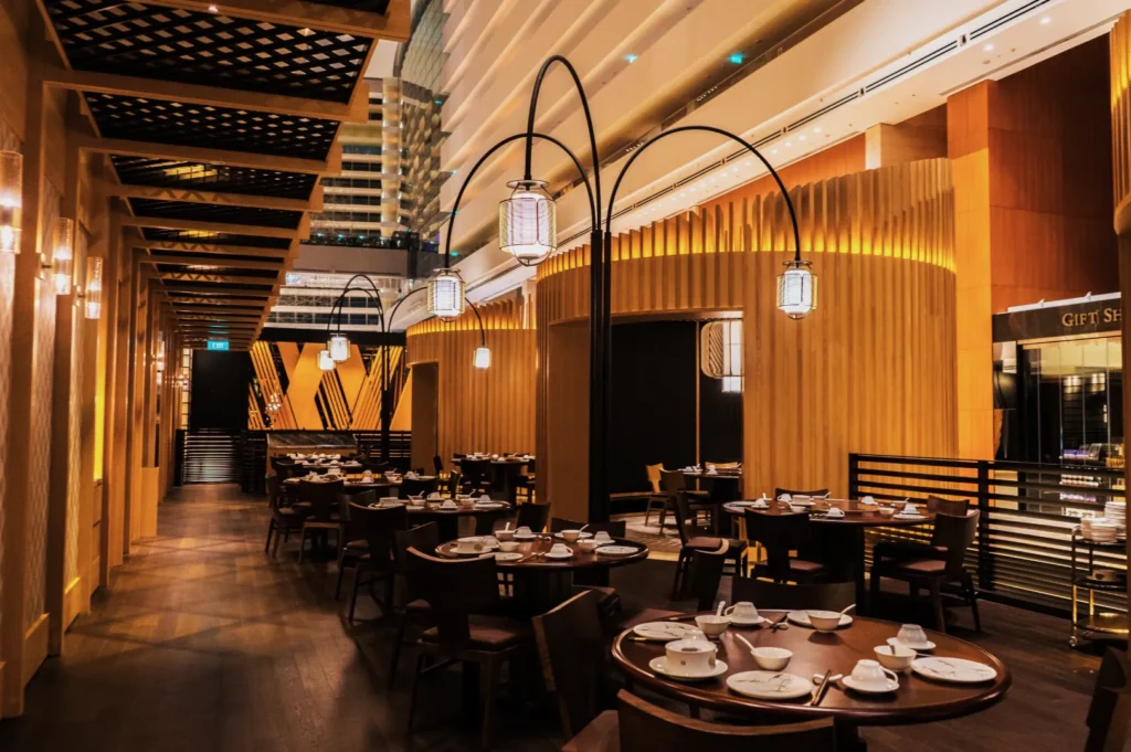 Elegant restaurant interior with warm lighting and modern decor. Dark wooden tables are set with white dishes. Vaulted ceiling and stylish lamps enhance the upscale ambiance.