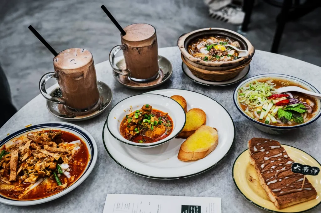 A table with diverse Asian dishes, including noodle soup, spicy stir-fry, and stews, alongside two chocolate drinks and toasted bread, creating a warm, hearty meal setting.