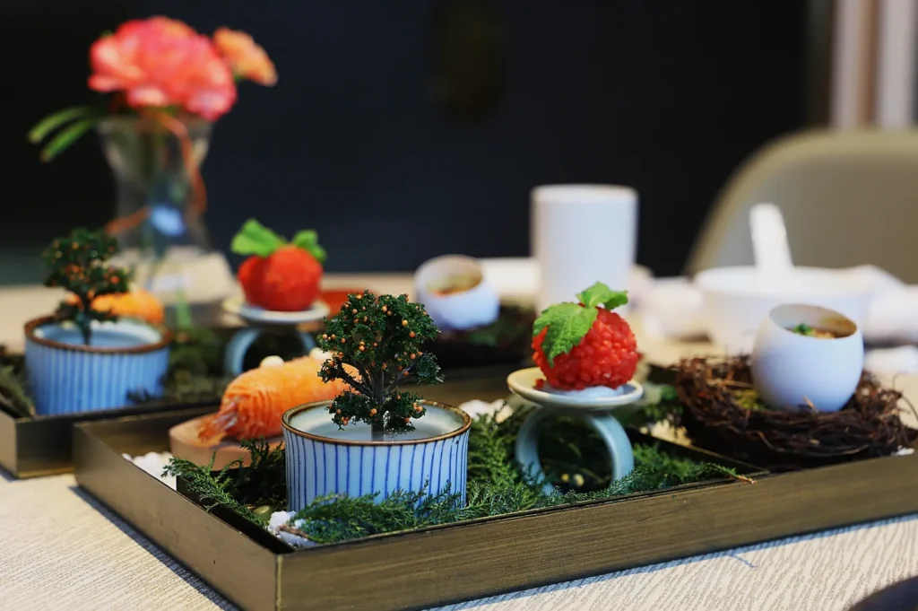 A beautifully arranged platter features intricate bonsai trees, gourmet dishes on pedestals, and a white egg in a nest, creating an artistic, elegant dining setting.