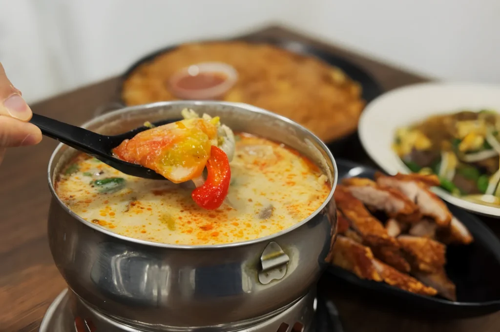 A steaming pot of creamy tom yum soup with a ladle lifting vegetables and shrimp, surrounded by crispy fried pork and vegetable stir-fry on a wooden table.