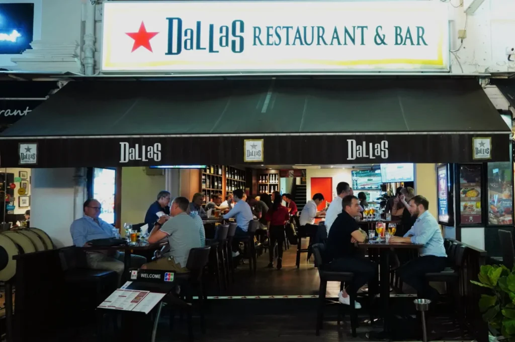 Front view of Dallas Restaurant & Bar, with patrons seated at tables under a canopy. The atmosphere is lively, with people dining and conversing.