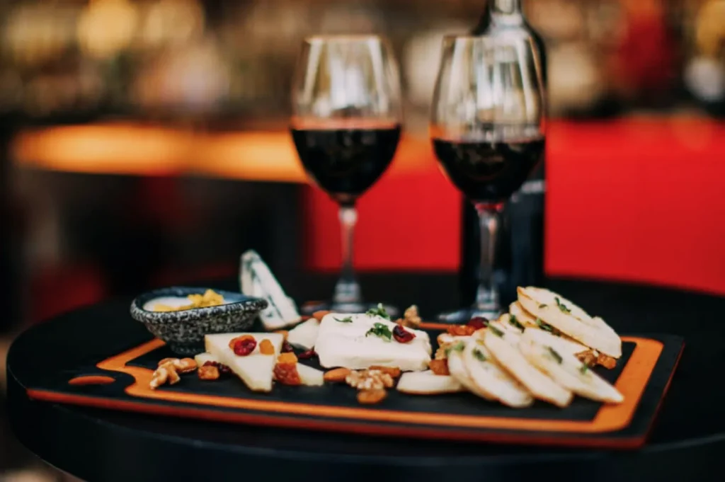 A cheese platter with sliced cheese, nuts, and dried fruits on a table, accompanied by two glasses of red wine. The setting is elegant and warm.