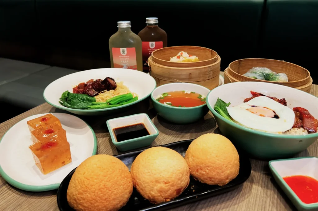 A variety of Asian dishes on a table, including dumplings, buns, noodles with roasted meat, and a bowl topped with a fried egg. Each dish is neatly presented, accompanied by dipping sauces and two iced tea bottles. The setting is casual and inviting.