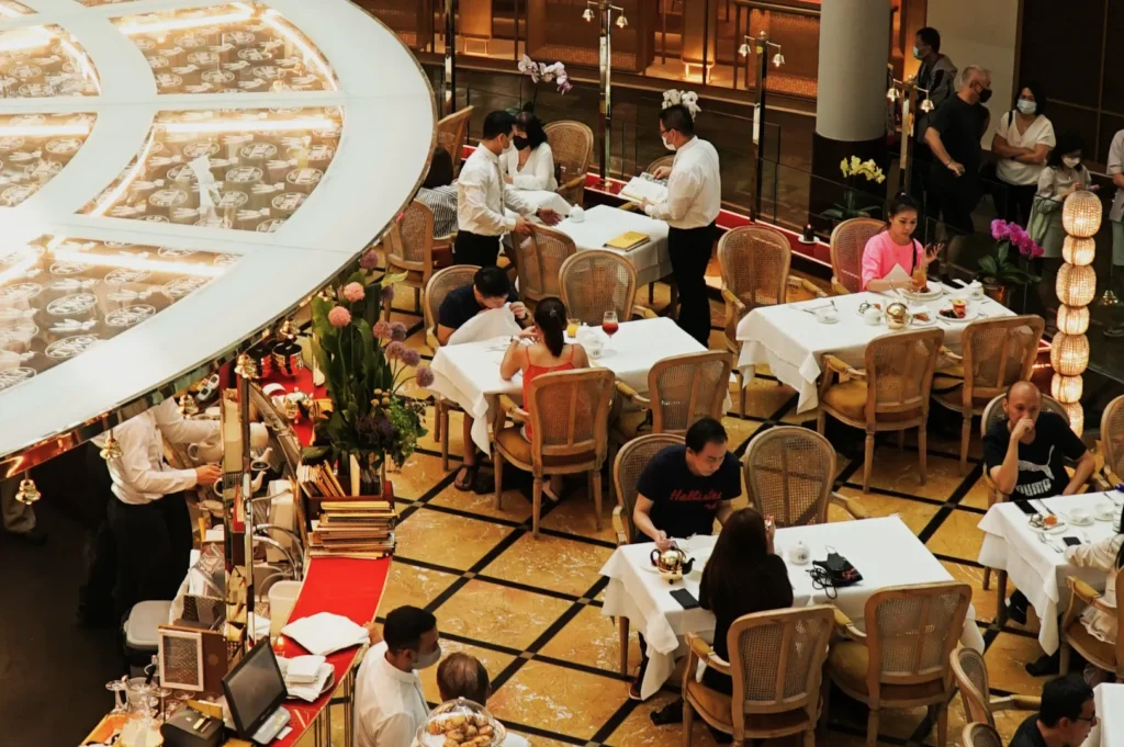 Overhead view of a busy restaurant with elegantly dressed diners seated at white-clothed tables. Waitstaff in white attire attend to guests in a warm, lively atmosphere.
