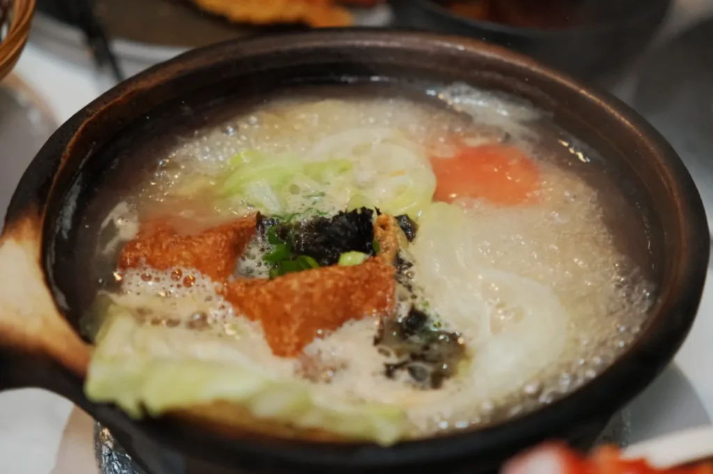 A steaming clay pot filled with bubbling broth, featuring vibrant cabbage, crispy tofu, seaweed, and a slice of tomato. Cozy and inviting atmosphere.