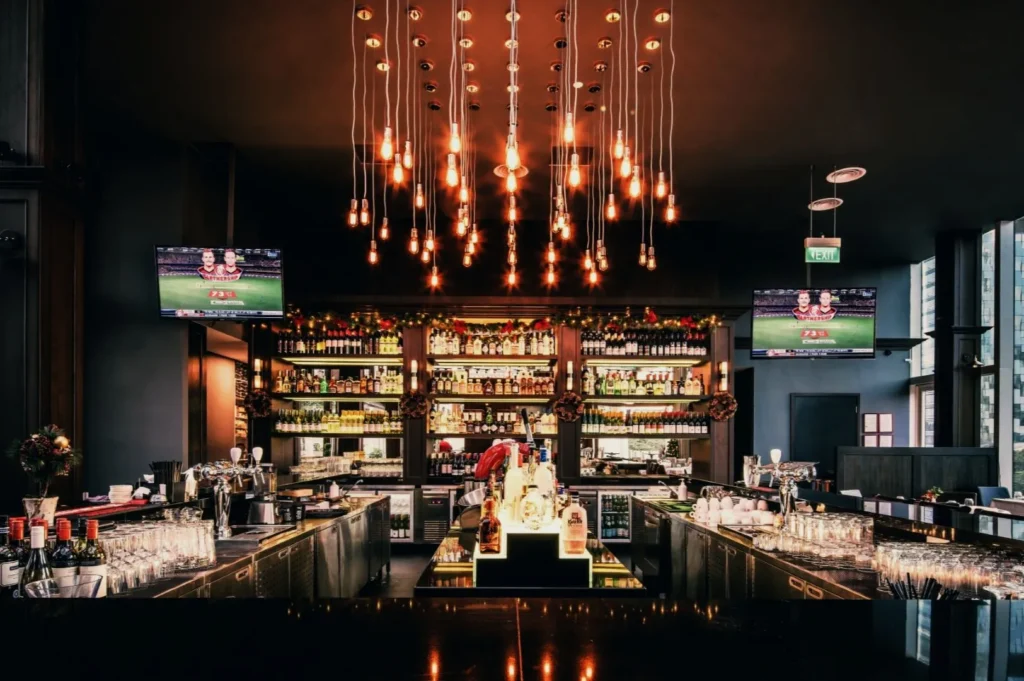 Elegant bar interior with warm lighting and sleek countertops. Bottles line the back shelves, and TV screens display a sports game, enhancing the lively atmosphere
