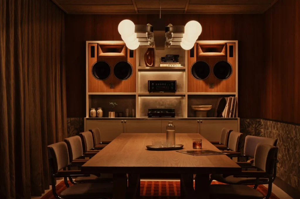 A cozy meeting room with a long wooden table and cushioned chairs. The wall features retro speakers, books, and warm lighting, creating a nostalgic ambiance.