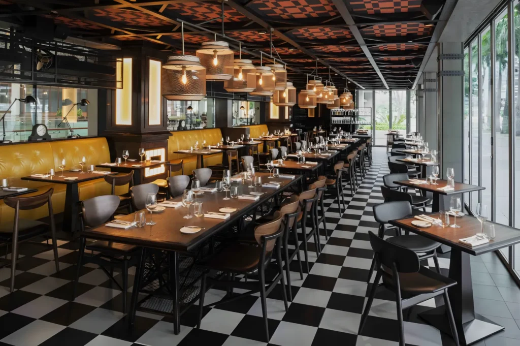 Modern restaurant with black-and-white checkered floor, yellow booths, brown wooden tables, and stylish hanging lights. Floor-to-ceiling windows brighten space.