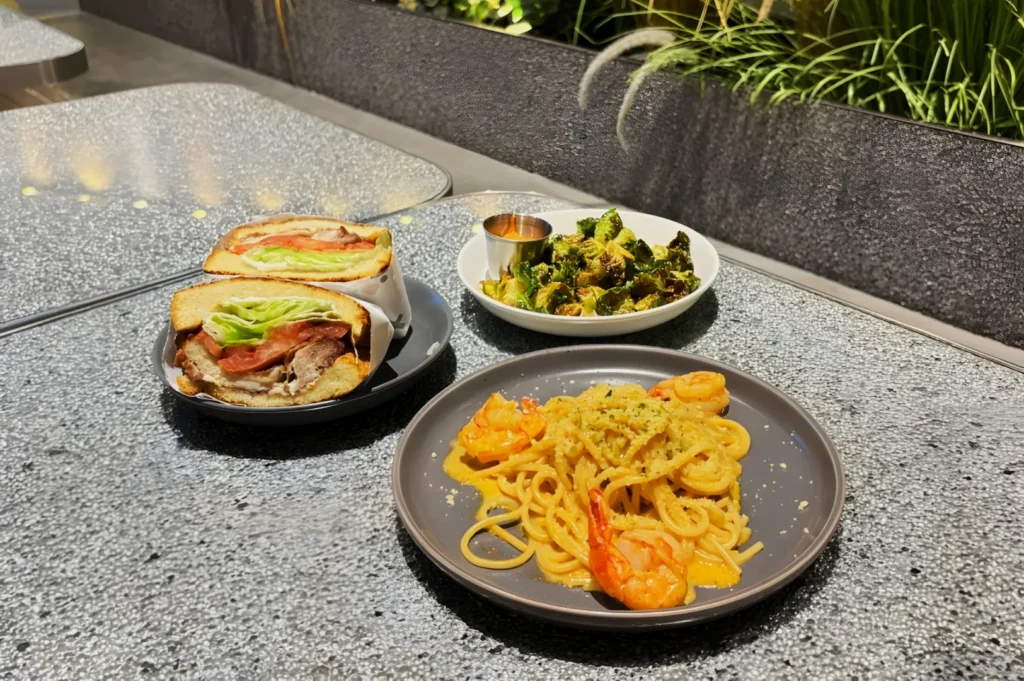 A table holds shrimp pasta on a gray plate, a sandwich filled with lettuce and tomato, and roasted broccoli with a dipping sauce. The setting is modern and well-lit.