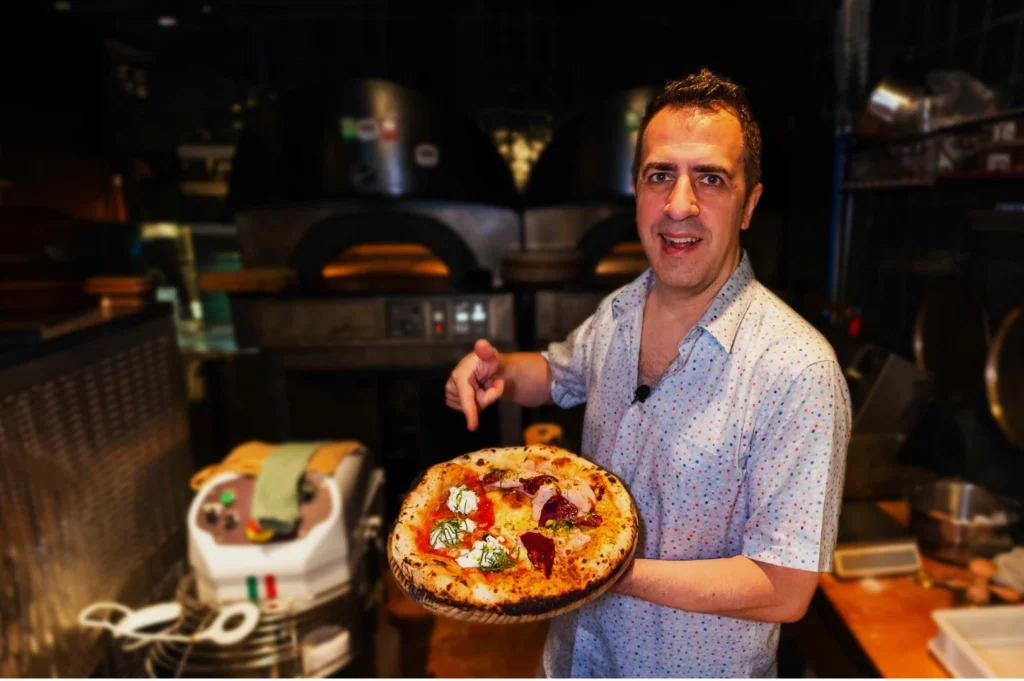 Chef Antonio Miscellaneo in a patterned shirt happily presents a freshly baked pizza topped with basil and ricotta in a cozy pizzeria kitchen, exuding a warm and inviting atmosphere