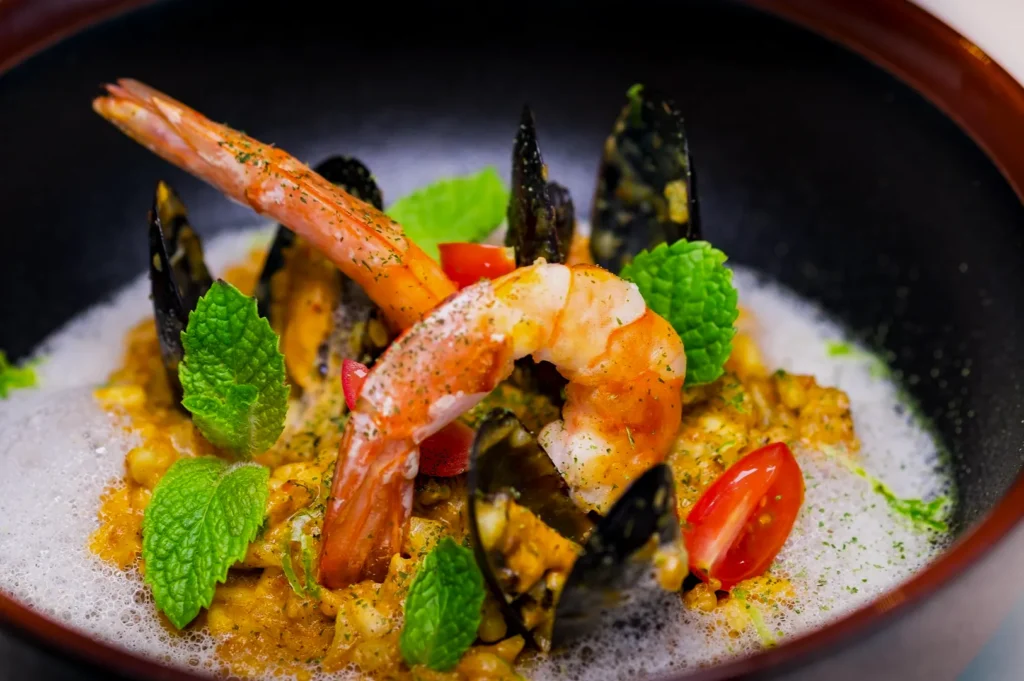A gourmet seafood dish with a large shrimp, mussels, cherry tomatoes, and fresh mint atop a creamy risotto, garnished with foam, in a black bowl.