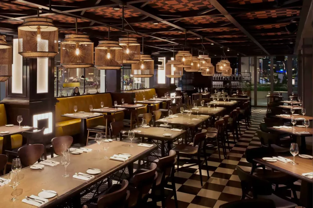 Cozy restaurant interior with wooden tables, elegant chairs, and warm lighting from hanging basket lamps. The floor features a black-and-white checkered pattern.