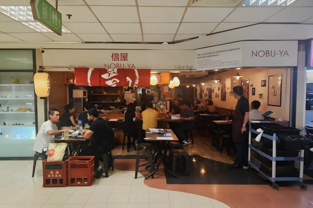 Cozy Japanese restaurant with red decor and dim lighting. Patrons sit at wooden tables, chatting and enjoying meals. A waiter stands nearby, adding to the lively atmosphere.