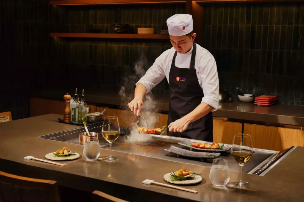 Chef in a white hat and black apron expertly grills food on a steaming teppan in a modern kitchen. Two wine glasses and plates set nearby.