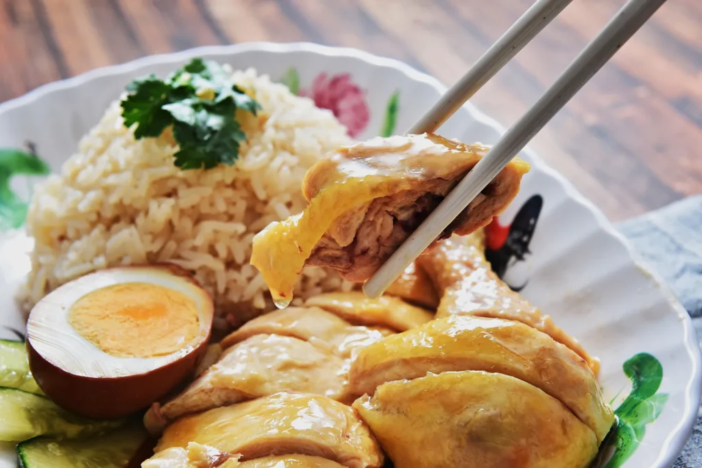 A plate of Hainanese chicken rice with sliced poached chicken, a halved boiled egg, cucumbers, and cilantro garnish. Chopsticks lift a chicken piece.