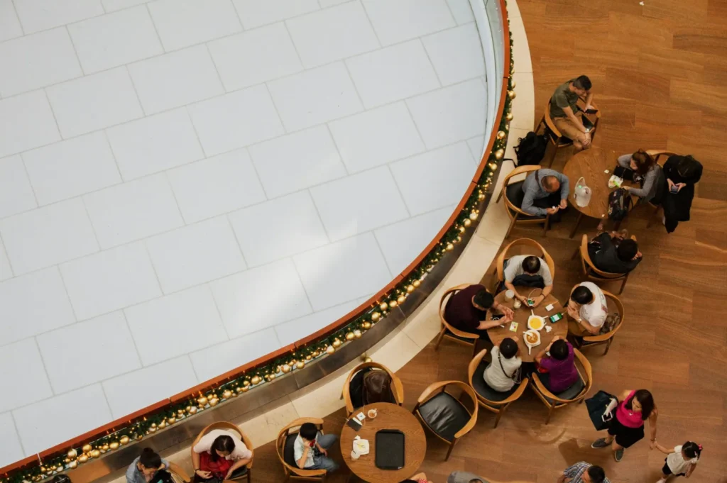 Overhead view of people seated at circular tables on wooden flooring by a large white circular area. The scene is lively and social.
