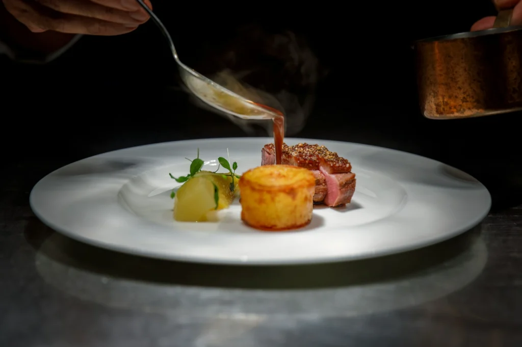 Chef artfully pours sauce over a plated dish featuring duck, a fondant potato, and garnish on a white plate, creating a refined and elegant presentation.