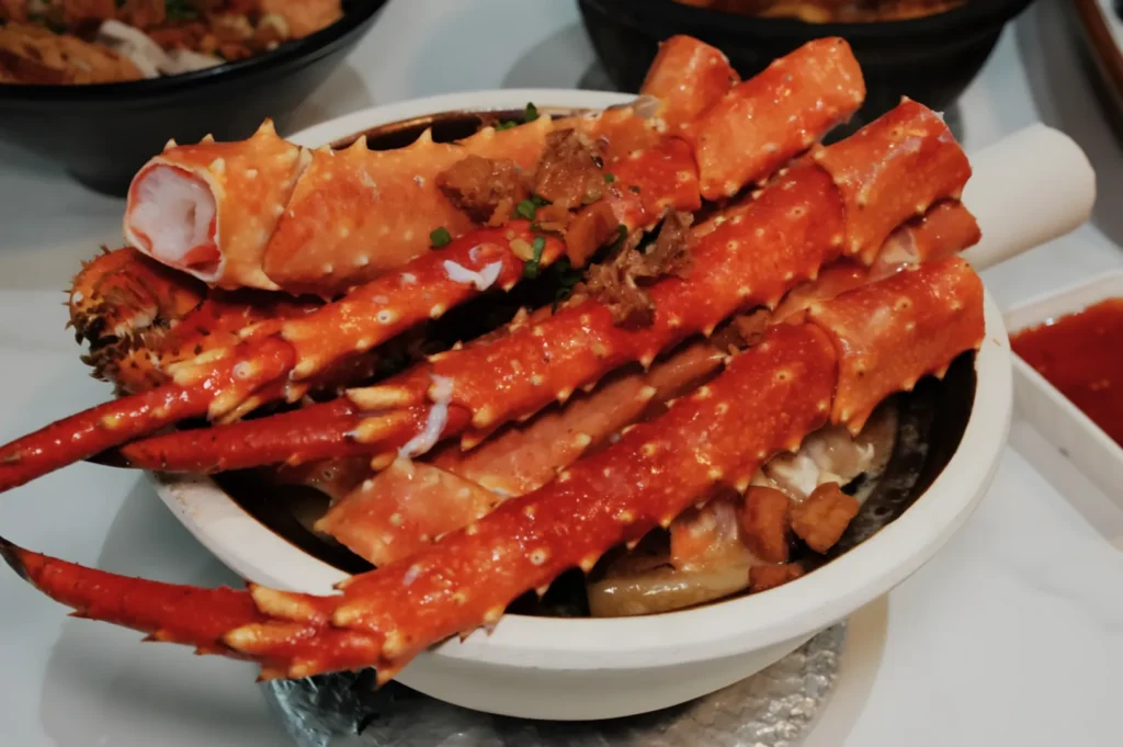 A dish of vibrant red crab legs rests atop a bowl filled with savory ingredients, garnished with green herbs and crumbled bits, creating an indulgent and appetizing presentation.