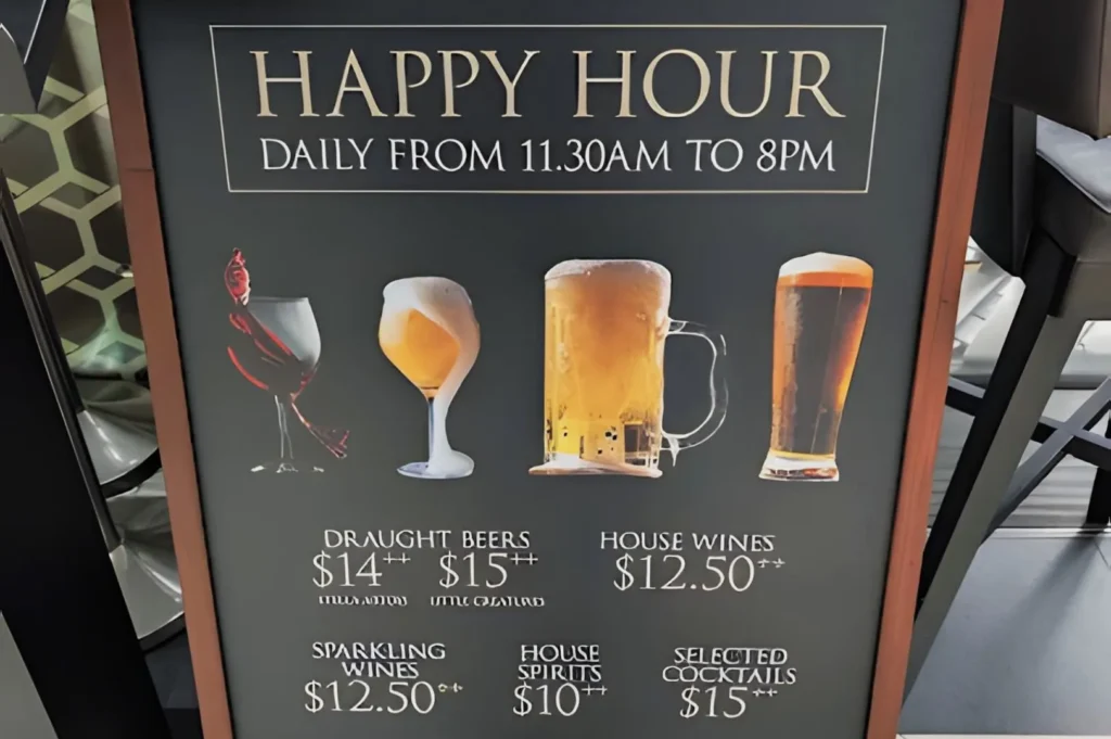 Chalkboard sign advertising Happy Hour from 11:30 AM to 8 PM with images of different drinks. Prices for beers, wines, spirits, and cocktails are displayed
