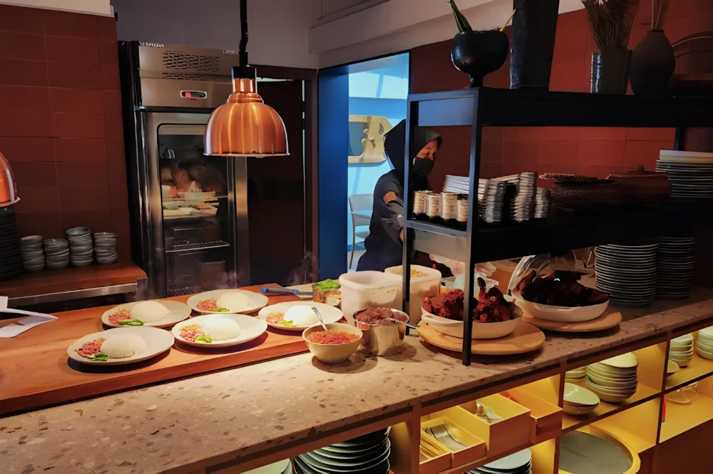 A restaurant kitchen scene with a counter serving plates of steamed rice, minced meat, and condiments. A person in the background is preparing food.