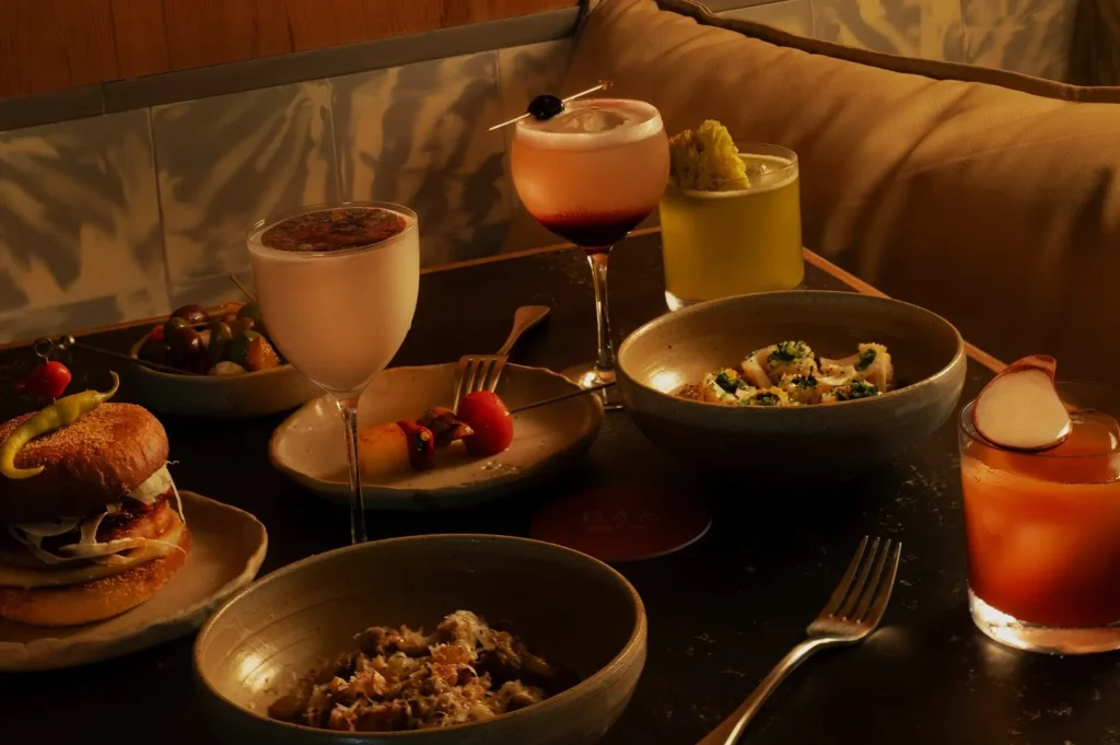 A cozy table setting features a variety of dishes including pasta, a sandwich, and skewers, accompanied by colorful cocktails and illuminated by warm lighting.
