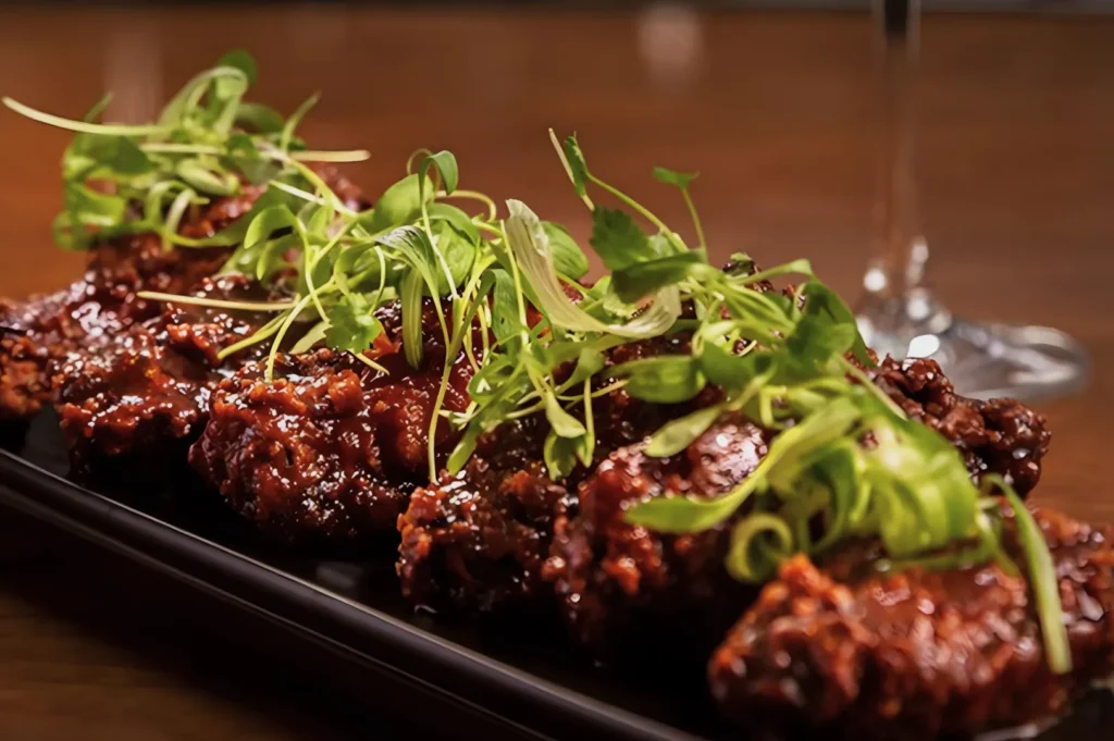 Close-up of crispy, glazed chicken wings on a rectangular black plate, garnished with fresh green herbs. The dish is set on a wooden table.