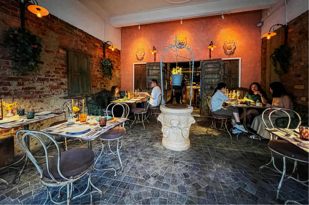 Cozy, rustic pizzeria with vintage decor, brick walls, and warm lighting. Patrons enjoy meals at elegant, wrought iron tables, creating a relaxed ambiance