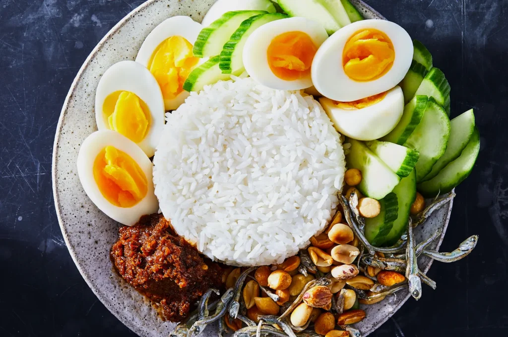 A plate of Nasi Lemak with rice, sliced cucumber, halved boiled eggs, sambal, roasted peanuts, and anchovies, arranged vibrantly for an appetizing look.