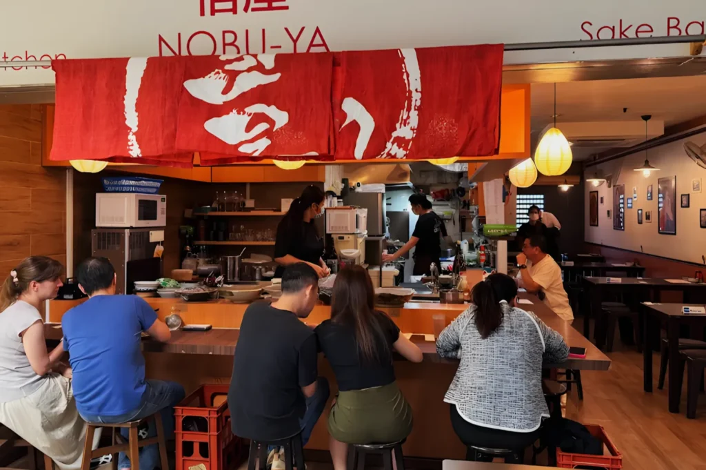A cozy Japanese restaurant interior with a red banner, customers seated at a wooden counter, chefs in the background preparing food, and warm lighting.