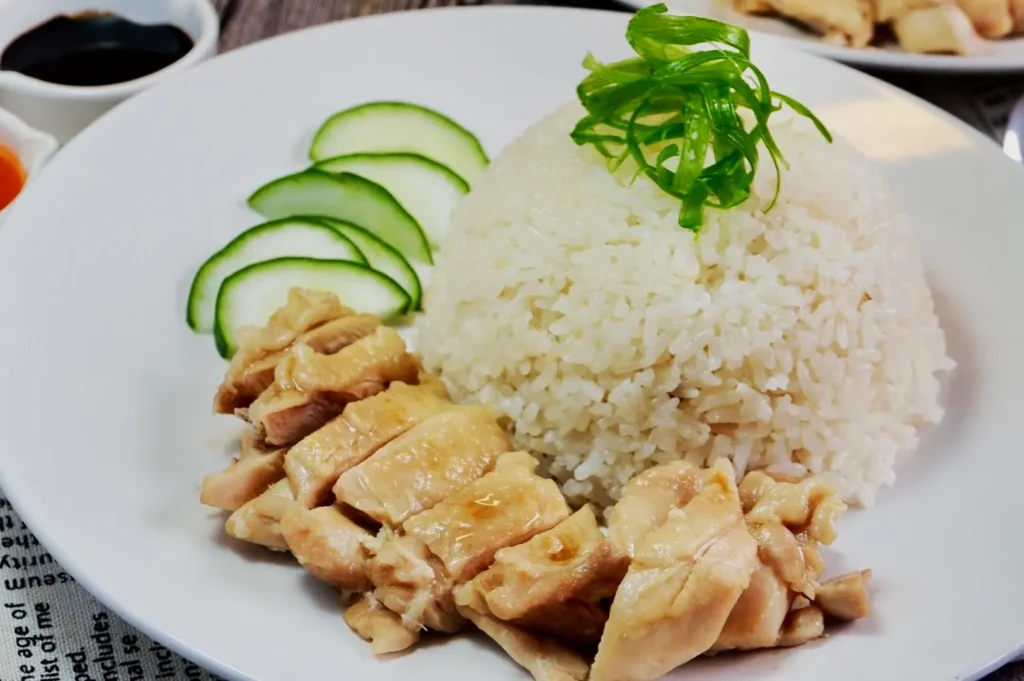 A plate of Hainanese chicken rice with tender sliced chicken, garnished with cucumber slices and green onions, next to a mound of white rice. Soy sauce dish visible.