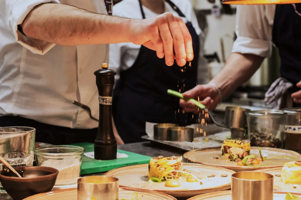 Chef in a white coat artfully seasons dishes in a busy kitchen, creating a vibrant, professional atmosphere. Plates showcase colorful, gourmet food.