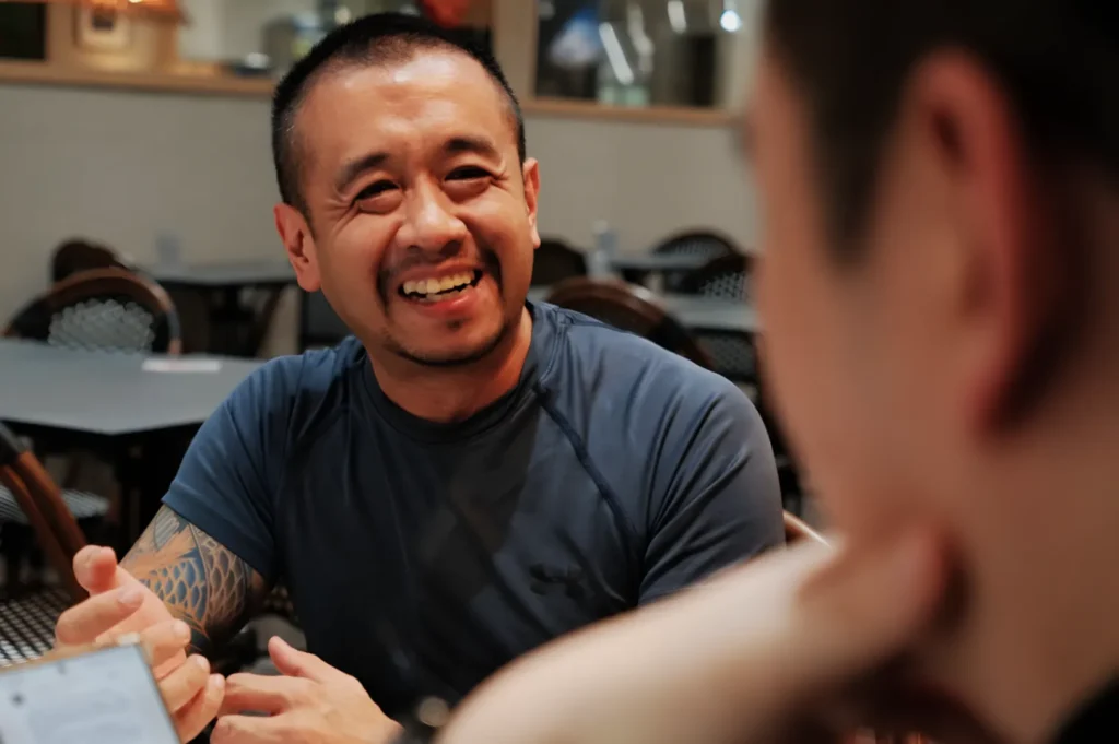 A smiling man with a short haircut and tattooed arm sits in a cafe, engaged in conversation. The atmosphere is warm and friendly, with chairs and tables in the background.