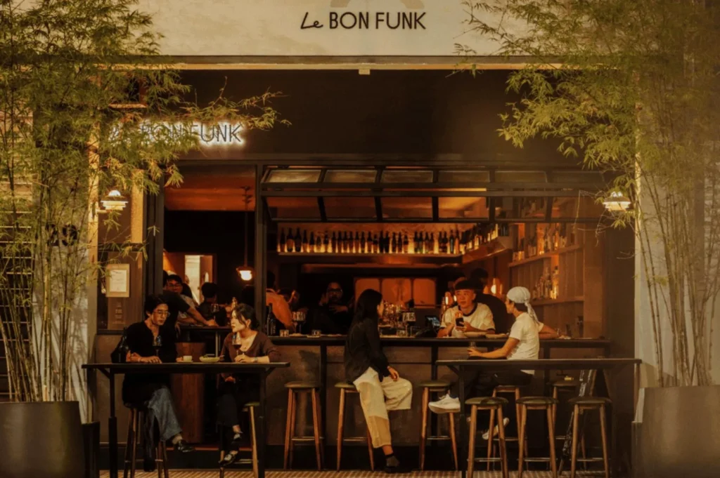 Exterior of a cozy café, "Le Bon Funk," with warm lighting. People are seated at tables, chatting and enjoying drinks, surrounded by greenery.