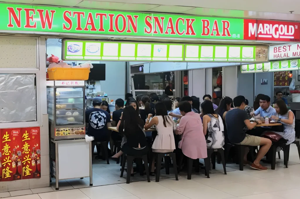 A bustling snack bar named "New Station Snack Bar" has people seated closely at wooden tables, enjoying meals. The ambiance is lively and social.