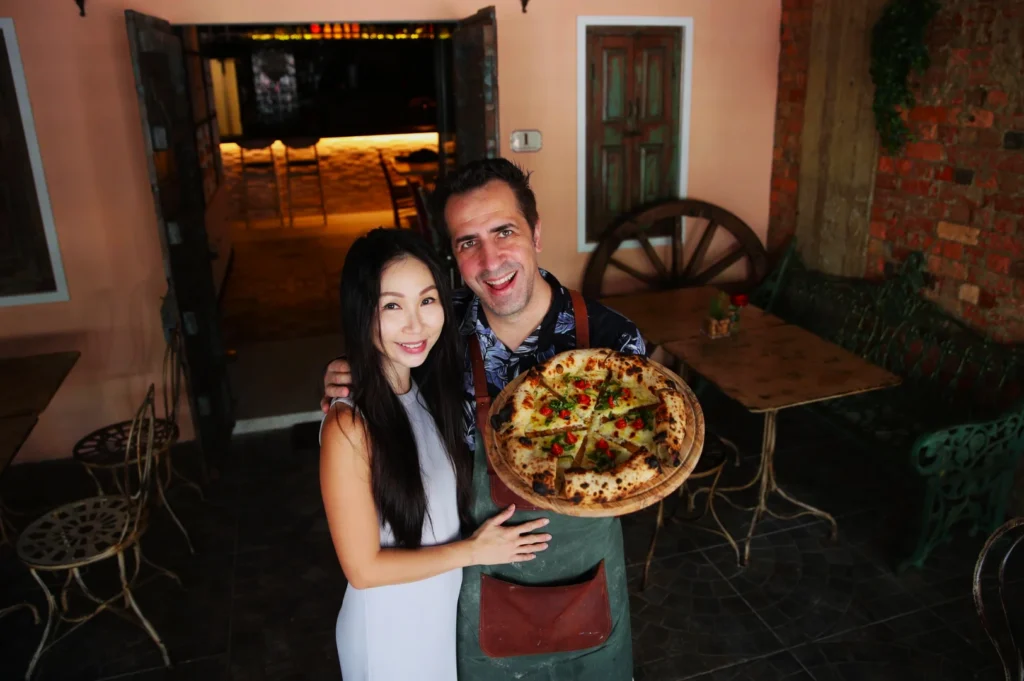 Chef Antonio Miscellaneo and his wife in a cozy, rustic pizzeria, holding a freshly-made pizza topped with herbs and tomatoes. Warm lighting enhances the inviting atmosphere.